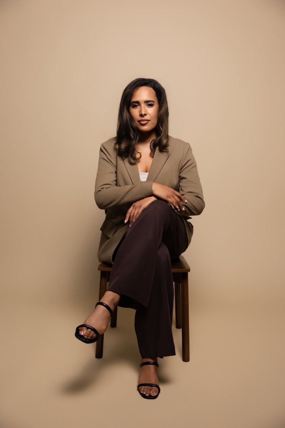 Camille (Cami) Boudreaux, relationship and life coach for women and founder of Fix Your Picker, sitting on a wooden chair with her legs crossed in a camel blazer and chocolate trousers, looking calmly at the camera