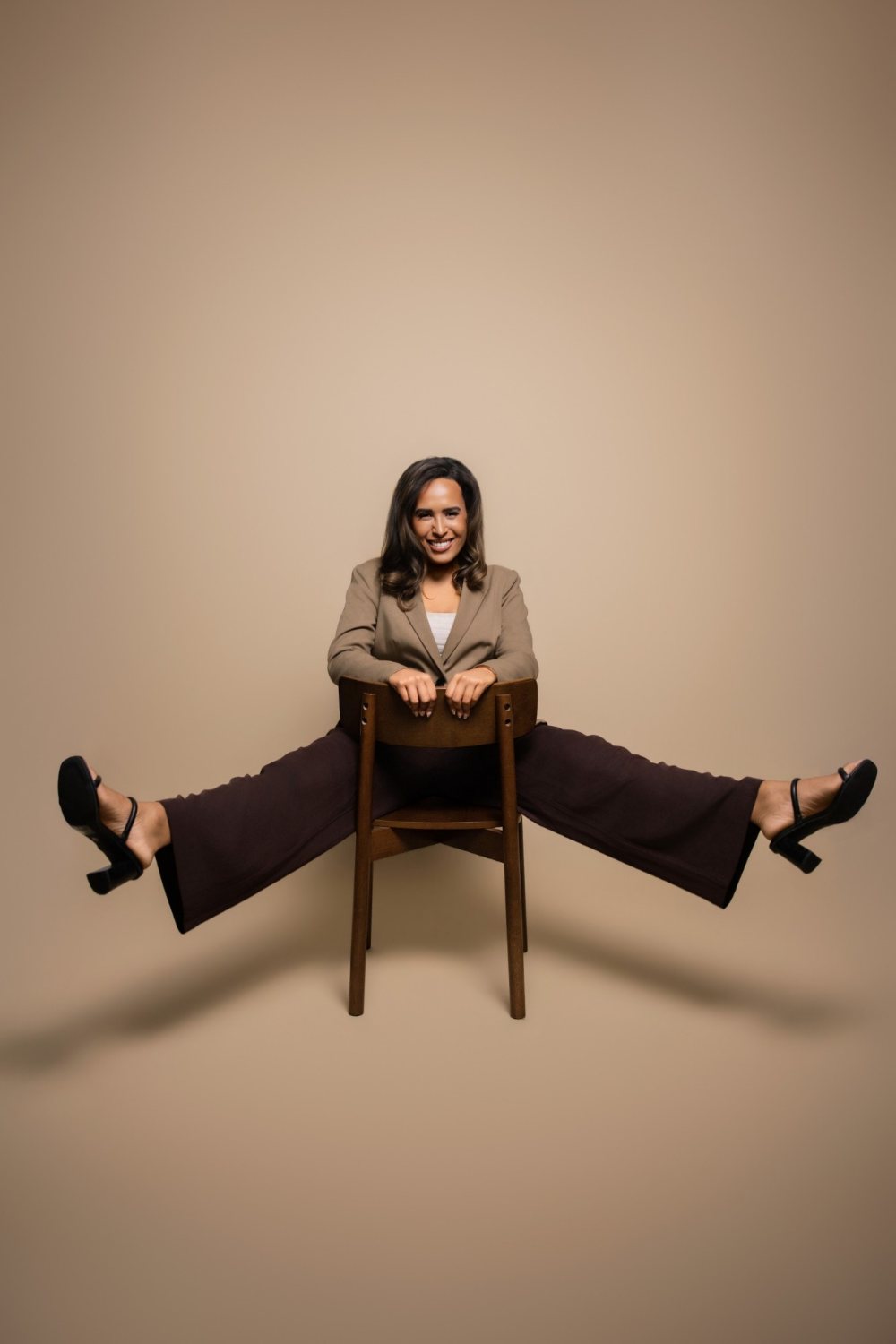 Camille (Cami) Boudreaux, relationship and life coach for women and founder of Fix Your Picker, laughing as she sits backwards on a wooden chair with her legs kicked out wide in a camel blazer
