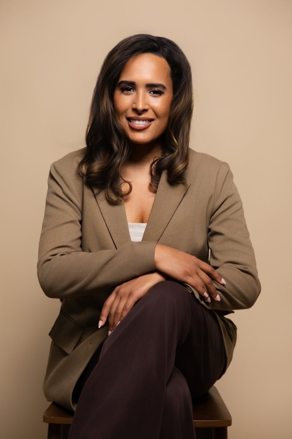 Camille (Cami) Boudreaux, relationship and life coach for women and founder of Fix Your Picker, smiling on a wooden chair in a camel blazer with her arms folded over her crossed knee